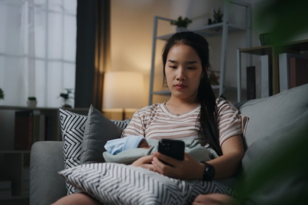 A young woman sits on a sofa in a dim room, holding a pillow on her lap. She is looking at a cell phone.