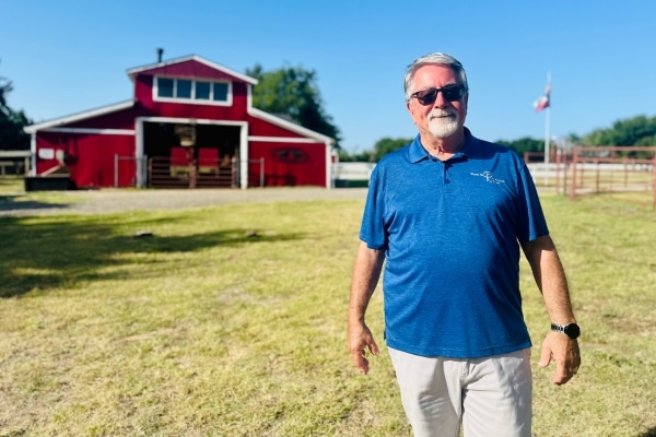 A man wearing a blue polo shirt stands in front of a red barn on a sunny day. He is smiling.