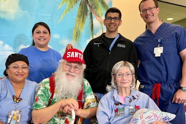 In front of a tropical backdrop, four people in medical scrubs smile at the camera. Seated in front, a bearded man in a baseball cap reading "Santa" and a woman in a blue shirt also smile for the photo.