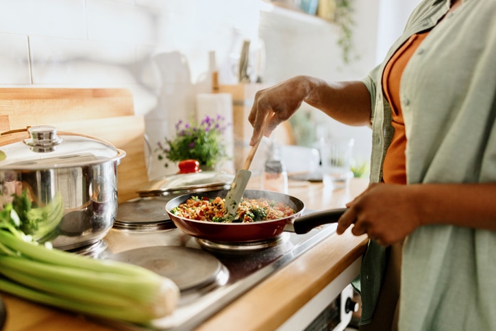 A woman stir-fries vegetables and rice. There's fresh celery in the foreground and a potted herb in the background.