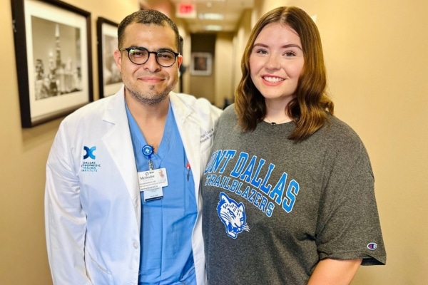 In a hospital hallway, a doctor stands beside a young woman wearing a UNT Dallas shirt. They are smiling.