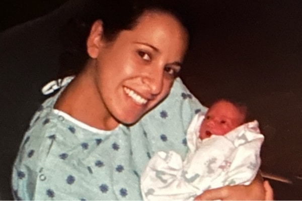 A woman wearing a medical gown holds an infant. She is smiling.