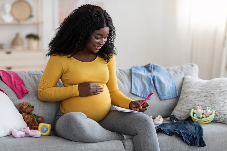 Happy pregnant woman sitting on sofa with notebook and pen, making birth plan.