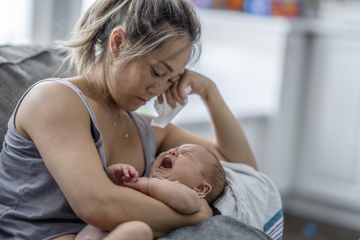 A middle-aged mother resting on her couch holds up her baby on her chest with a tired and stressed expression.