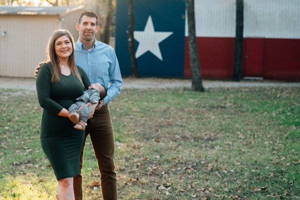 Jessica and Michael Farrah hold baby Miles with a building painted with a giant Texas flag in the background.