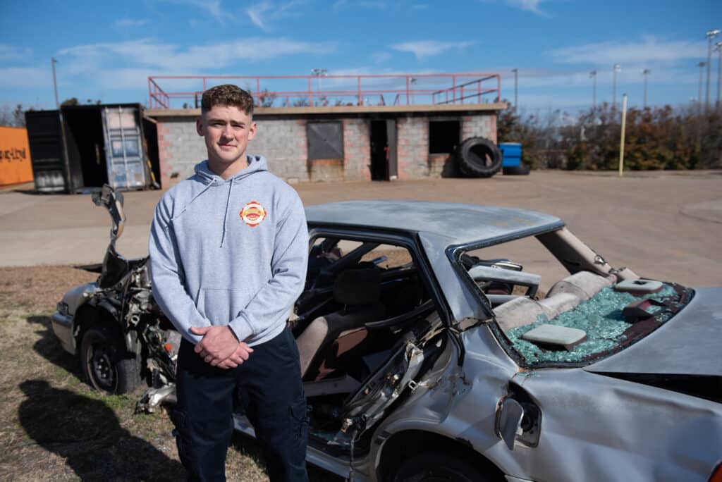 Young firefighter stands in front of wrecked car used for training.