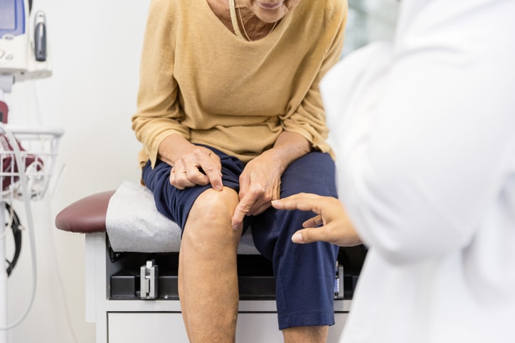 Unrecognizable senior woman, sitting on the examination table, shows the unrecognizable female doctor the exact place where her knee hurts.