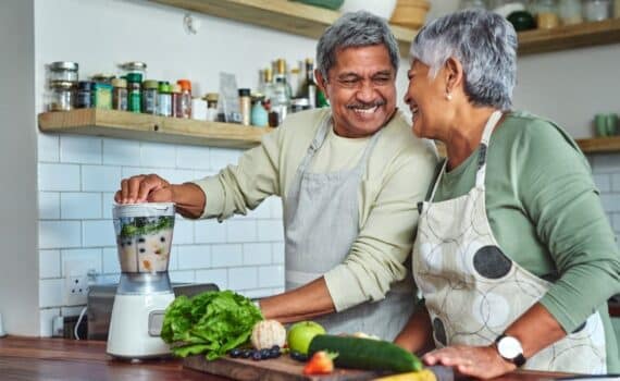 An older couple share a laugh as they prepare a smoothie in the kitchen.