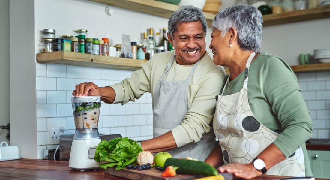 An older couple share a laugh as they prepare a smoothie in the kitchen.