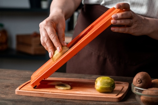 Close-up of unrecognizable woman cutting kiwi on a mandolin slicer above wooden board in kitchen