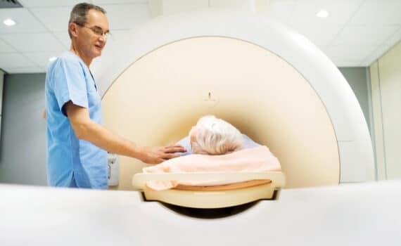 A patient undergoes an MRI test while a nurse comforts him with a hand on his shoulder.