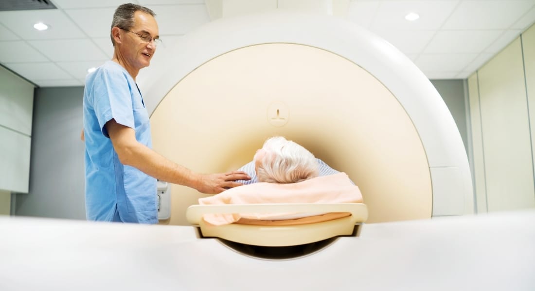 A patient undergoes an MRI test while a nurse comforts him with a hand on his shoulder.