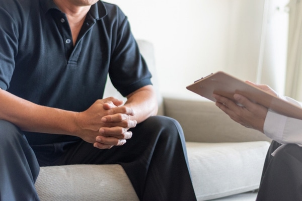 An unrecognizable man speaks to his doctor while sitting on a white couch.