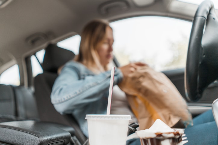 Woman sitting in her car, opening a take-out bag, eating fast food and drinking soda. In a rush, multi-tasking, eating take-out burger, french fries and dessert while driving a car.