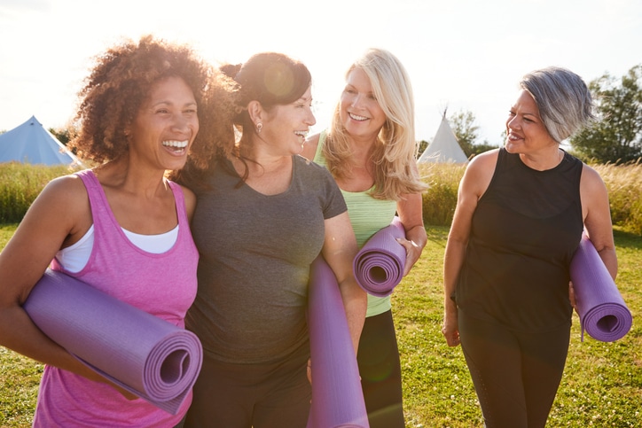 Group of mature female friends on outdoor yoga retreat walking along path through campsite