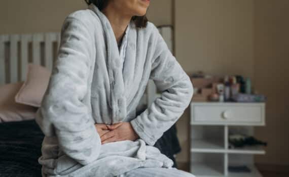 A woman in a bathrobe sits on her bed while suffering from abdominal pain.