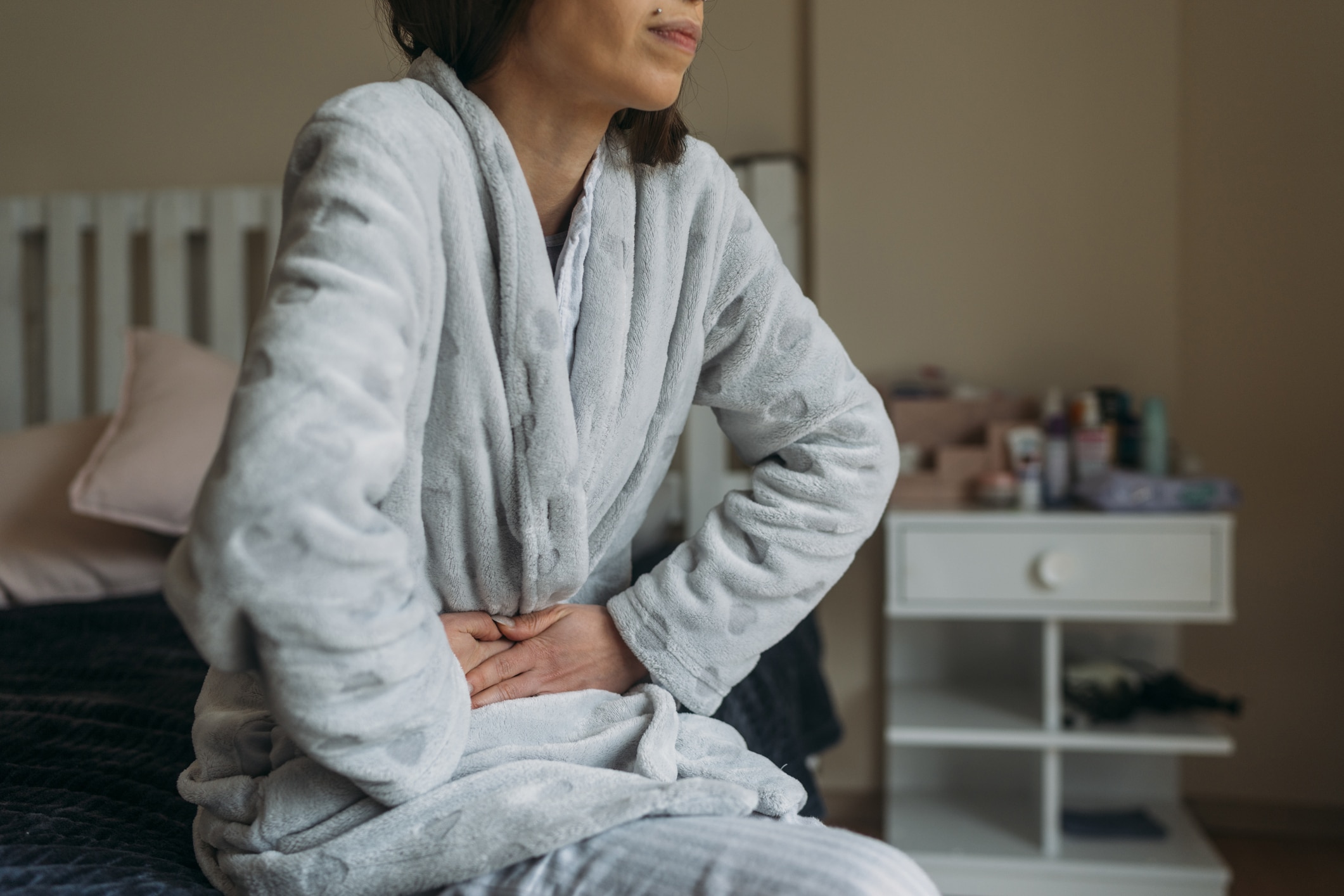 A woman in a bathrobe sits on her bed while suffering from abdominal pain.