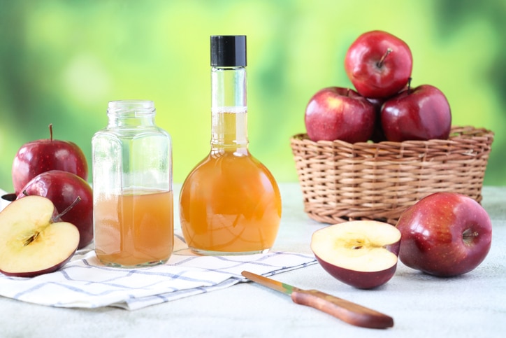 Stock photo showing close-up view of a wicker basket containing red apples besides two plastic bottles, one open and one closed containing apple cider vinegar, against a mottled green background. Apple cider vinegar is thought to have several health benefits including weight loss, lower cholesterol, whiten teeth and improve acne. Healthy lifestyle concept.