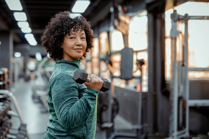 A Middle-aged, Plus-size Woman Smiling at the Camera in the Gym. She Exudes Confidence and Happiness, Reflecting a Sense of Accomplishment. Her Comfortable Activewear Fits Well, and the Gym Environment With Various Exercise Equipment in the Background Highlights Her Dedication to Her Fitness Journey. This Image Conveys a Positive Message of Resilience, Self-care, and the Importance of Maintaining Health at Any Age or Body Type.
