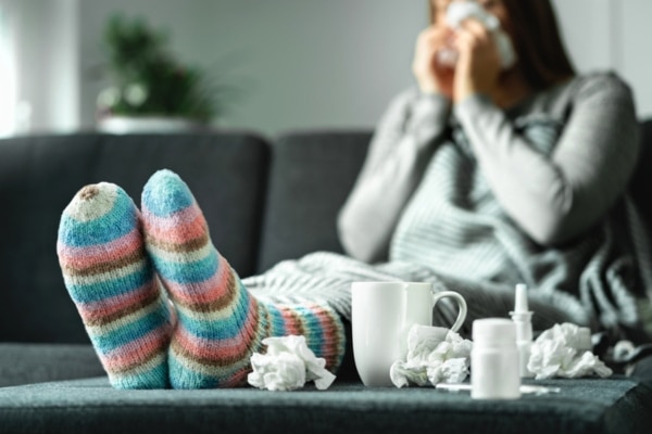 A woman blows her nose while sitting on her couch next to a footstool piled with spent tissues.