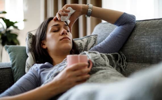 A woman lies on a couch with a cup of tea while suffering with the flu.