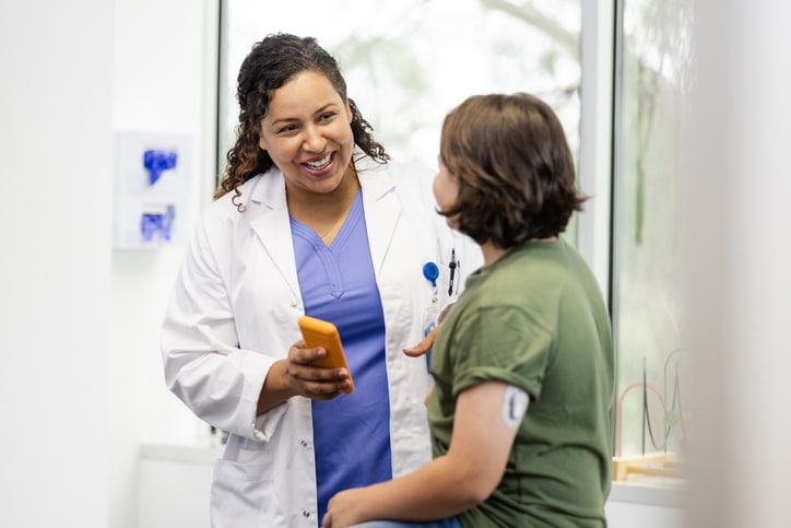 The mid adult female doctor smiles while talking to the young male patient to ask questions about the glucose monitor he has on his arm.