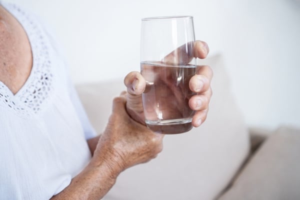 An older woman steadies her hand while holding a glass of water and suffering a Parkinson's tremor.