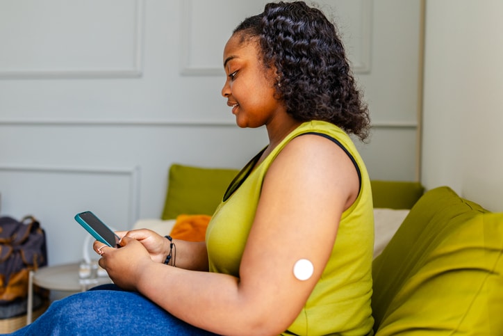 Sitting on a green couch in a cozy room, a young black woman monitors her type 1 diabetes using her phone, emphasizing the importance of health management in everyday life.