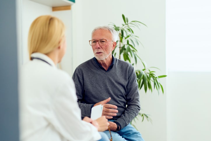 Elderly man speaks with a female doctor during a thoughtful medical consultation in a bright clinic, conveying trust, concern, and professional care for senior health and patient-doctor communication.