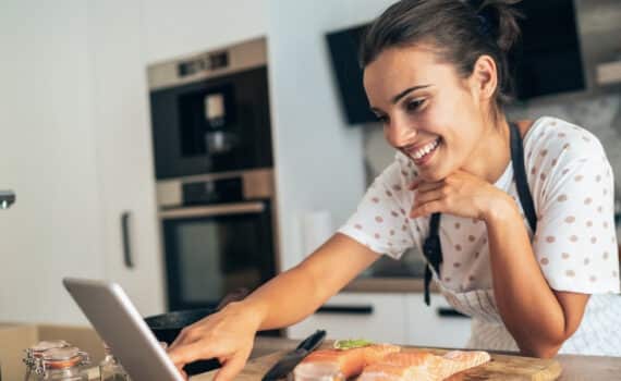 Woman looks at tablet smiling and reading along while in the kitchen preparing some salmon to cook, used to describe the relationship between certain foods and fertility