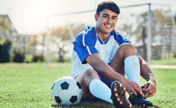A soccer player sits next to a ball tying his cleats with a goal in the background.