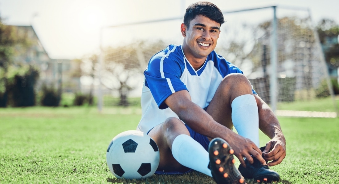 A soccer player sits next to a ball tying his cleats with a goal in the background.