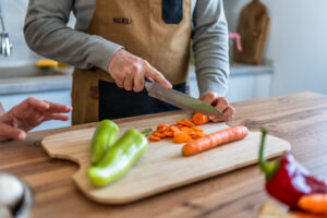 Close-up of a mature man wearing an apron, skillfully slicing carrots on a wooden cutting board, with fresh peppers nearby, in a brightly lit kitchen