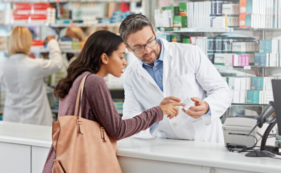 A pharmacist looks at a box of medicine with a customer.