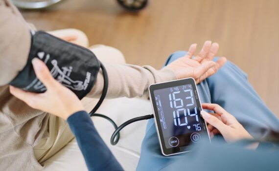 Close up woman caregiver nurse wearing blue scrubs checks blood pressure monitor and heart rate monitor with digital pressure gauge to elderly woman retirement sitting on bed in bedroom,