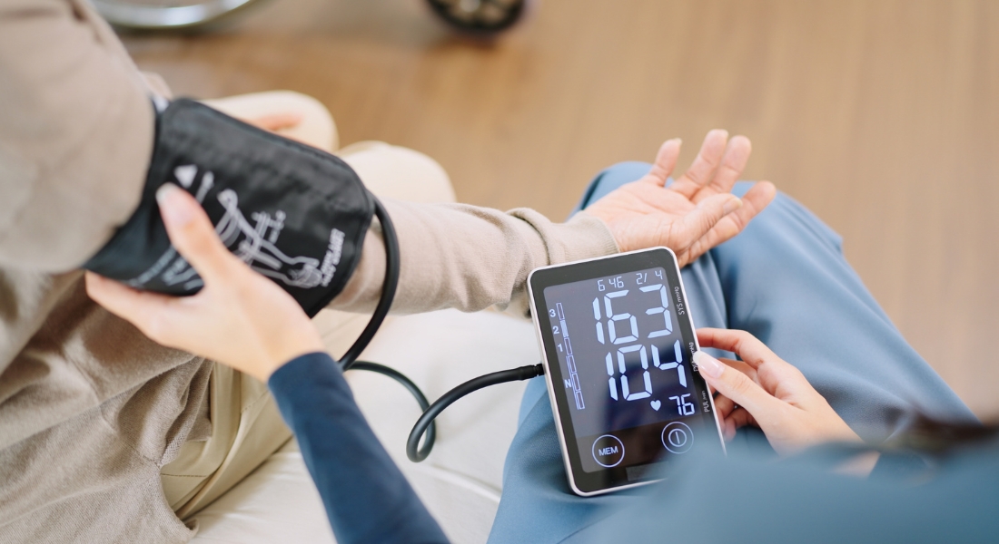 Close up woman caregiver nurse wearing blue scrubs checks blood pressure monitor and heart rate monitor with digital pressure gauge to elderly woman retirement sitting on bed in bedroom,